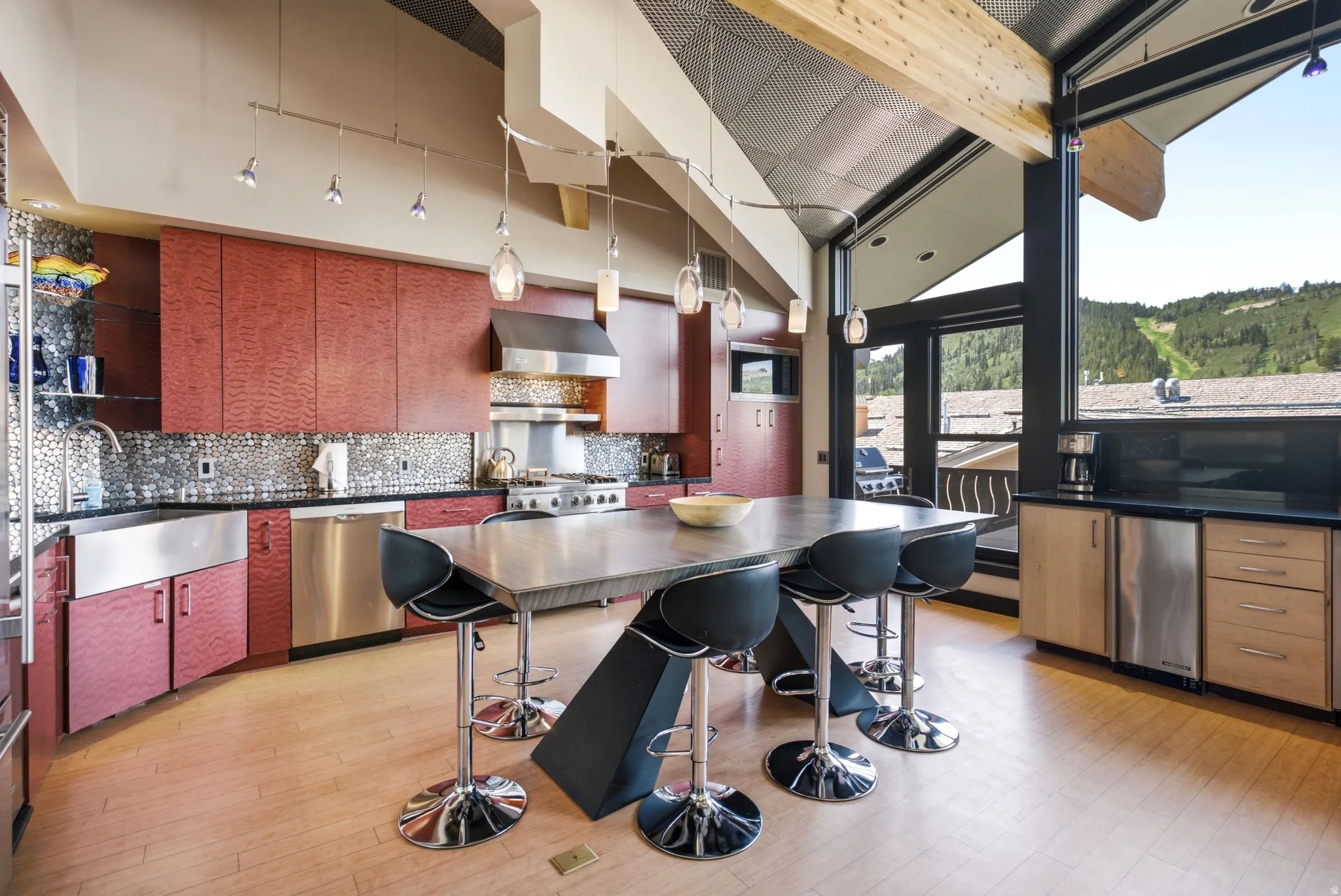 Modern kitchen featuring a large central island, stainless steel appliances, red wood-finish cabinetry, a textured backsplash, and expansive windows