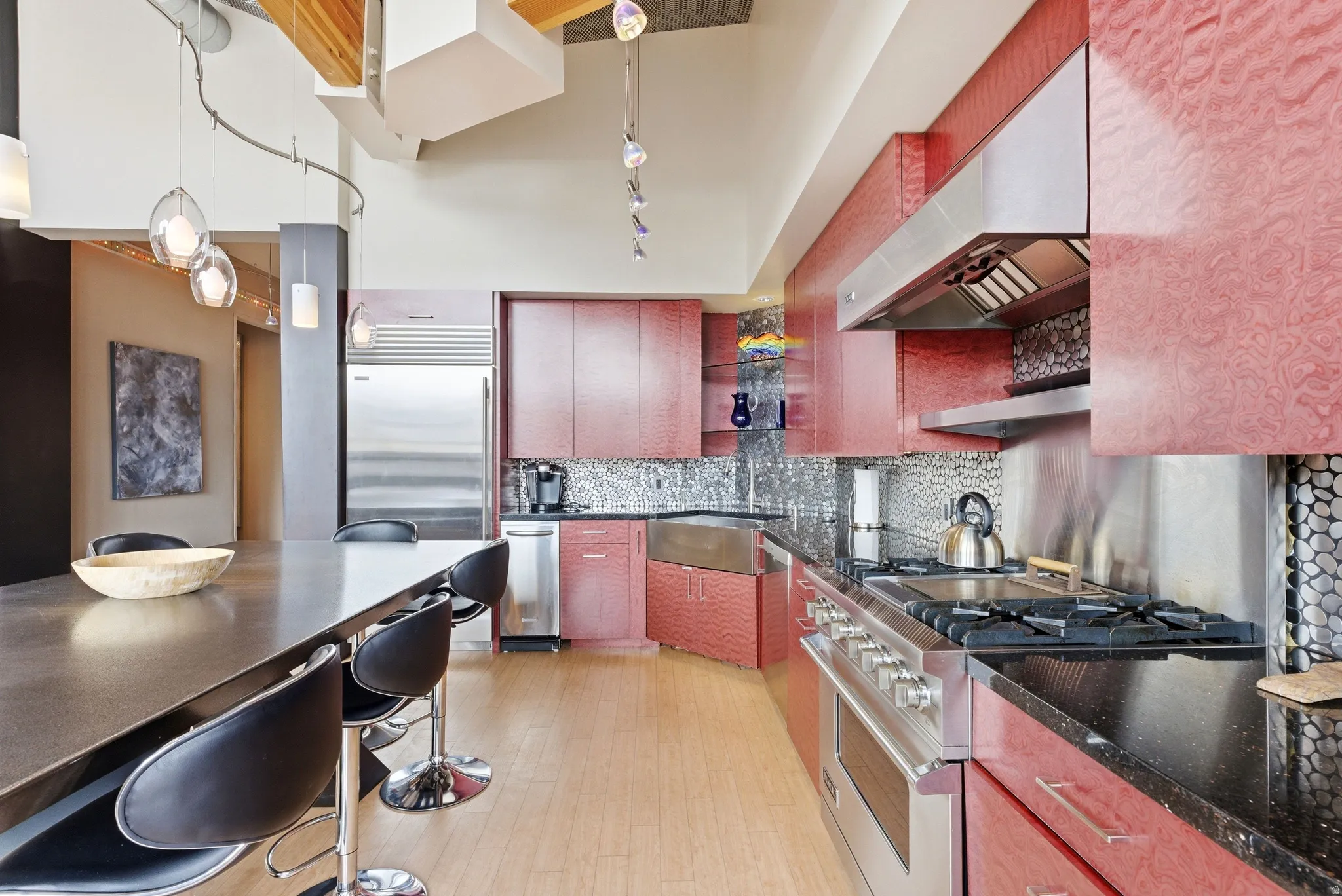 Modern kitchen featuring red textured cabinetry, stainless steel appliances, black countertops, and a stone pebble backsplash