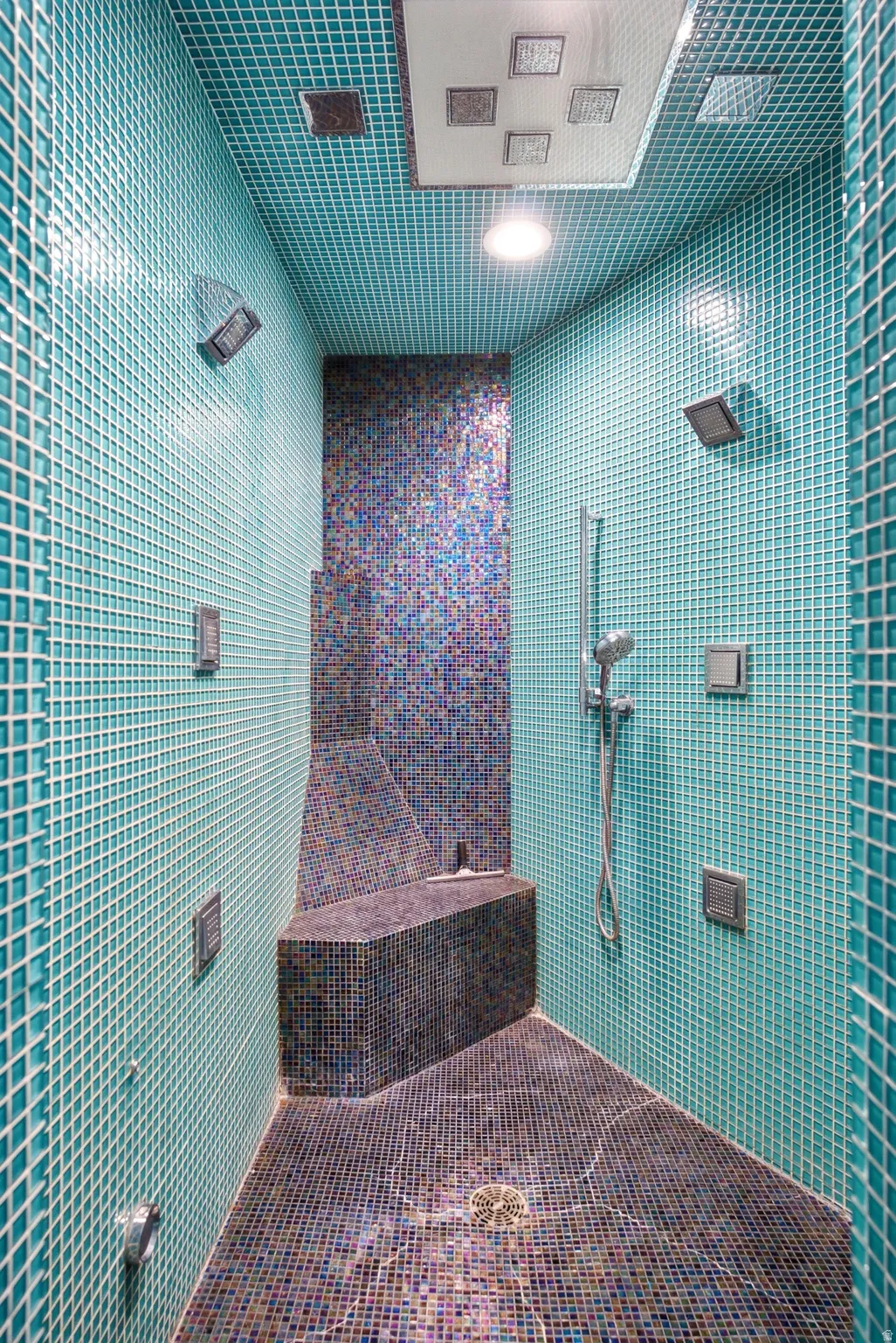 Custom-tiled shower featuring teal and multi-colored mosaic tiling, multiple wall-mounted sprayers, a built-in bench, a handheld showerhead, and an overhead rainfall shower panel