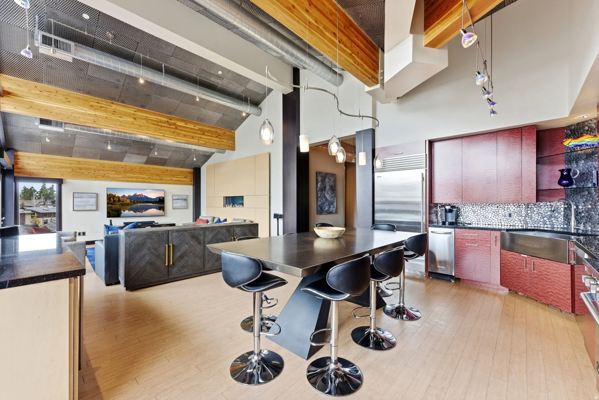 Open-concept living space featuring vaulted ceilings with exposed wood beams, light wood-finish flooring, a contemporary fireplace, a spacious kitchen island with seating, and stainless steel appliances