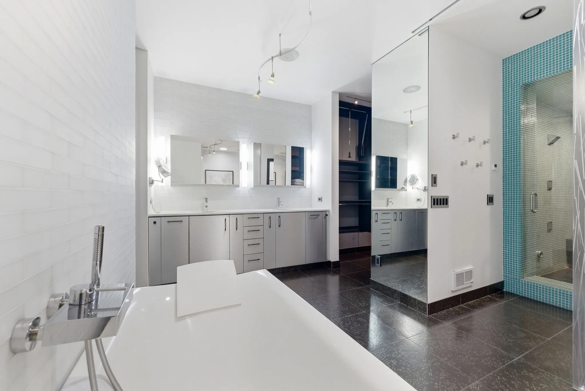 Freestanding soaking tub with chrome fixtures, dual vanities with undermount sinks, mirrored sliding door, built-in shelving, and a glass-enclosed shower with blue mosaic tile accents