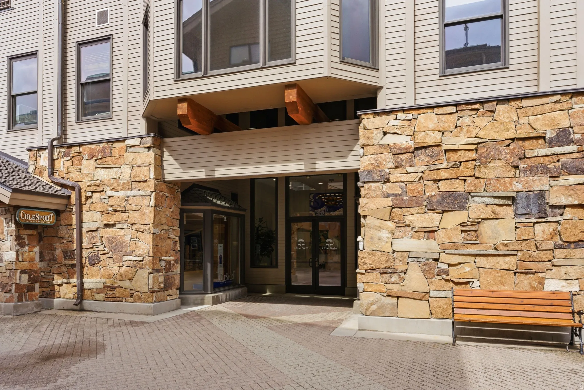 Building exterior featuring a stone and siding facade, a prominent entrance with glass double doors, a paved brick forecourt, and a built-in wooden bench