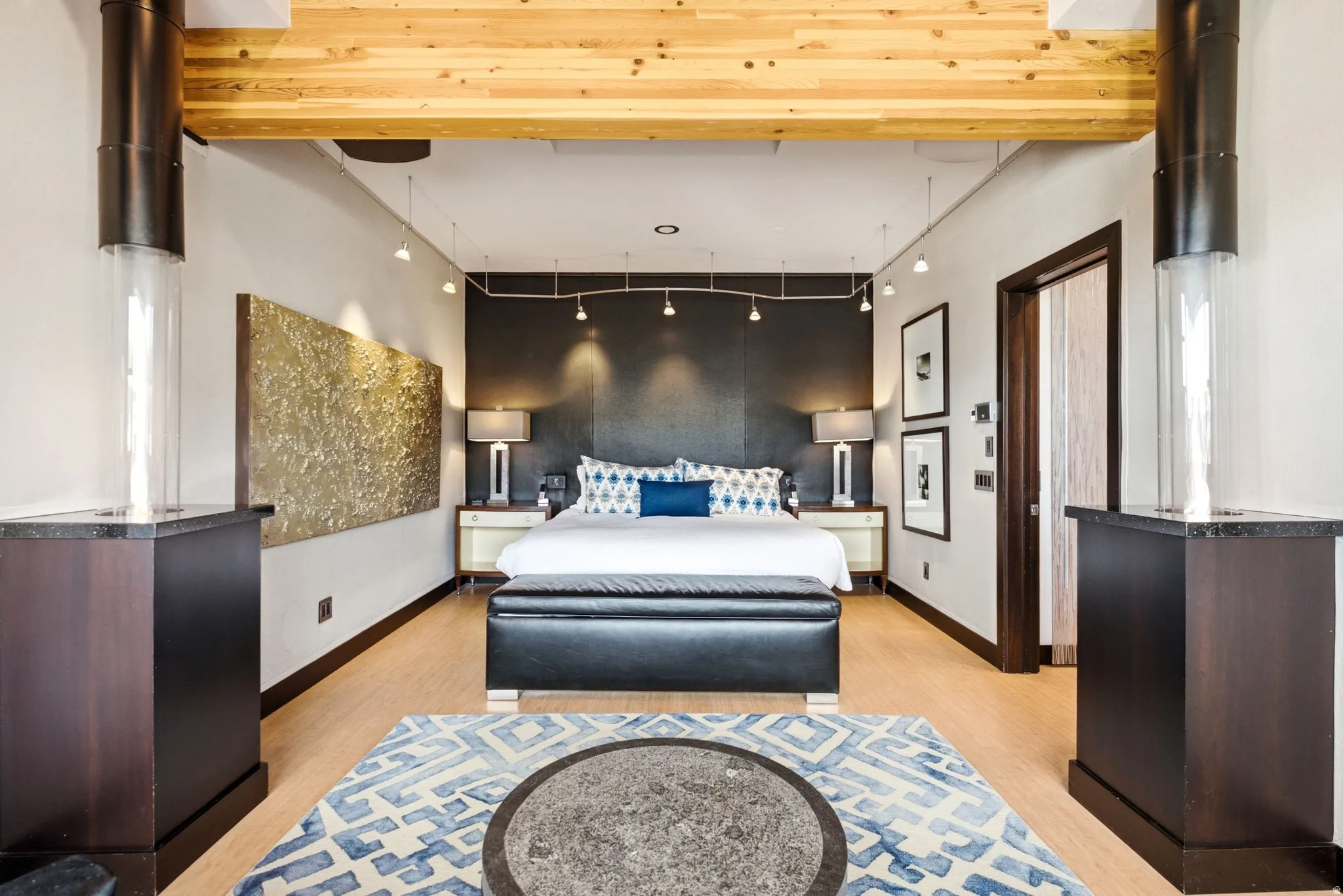 Spacious bedroom featuring light wood-finish flooring, exposed wood ceiling beams, and a dark accent wall