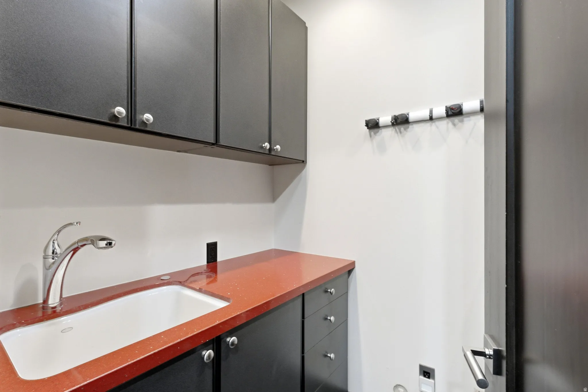 Laundry room featuring dark cabinetry, a built-in sink with chrome faucet, a vibrant countertop, and wall-mounted storage