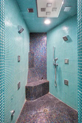 Custom-tiled shower featuring teal and multi-colored mosaic tiling, multiple wall-mounted sprayers, a built-in bench, a handheld showerhead, and an overhead rainfall shower panel