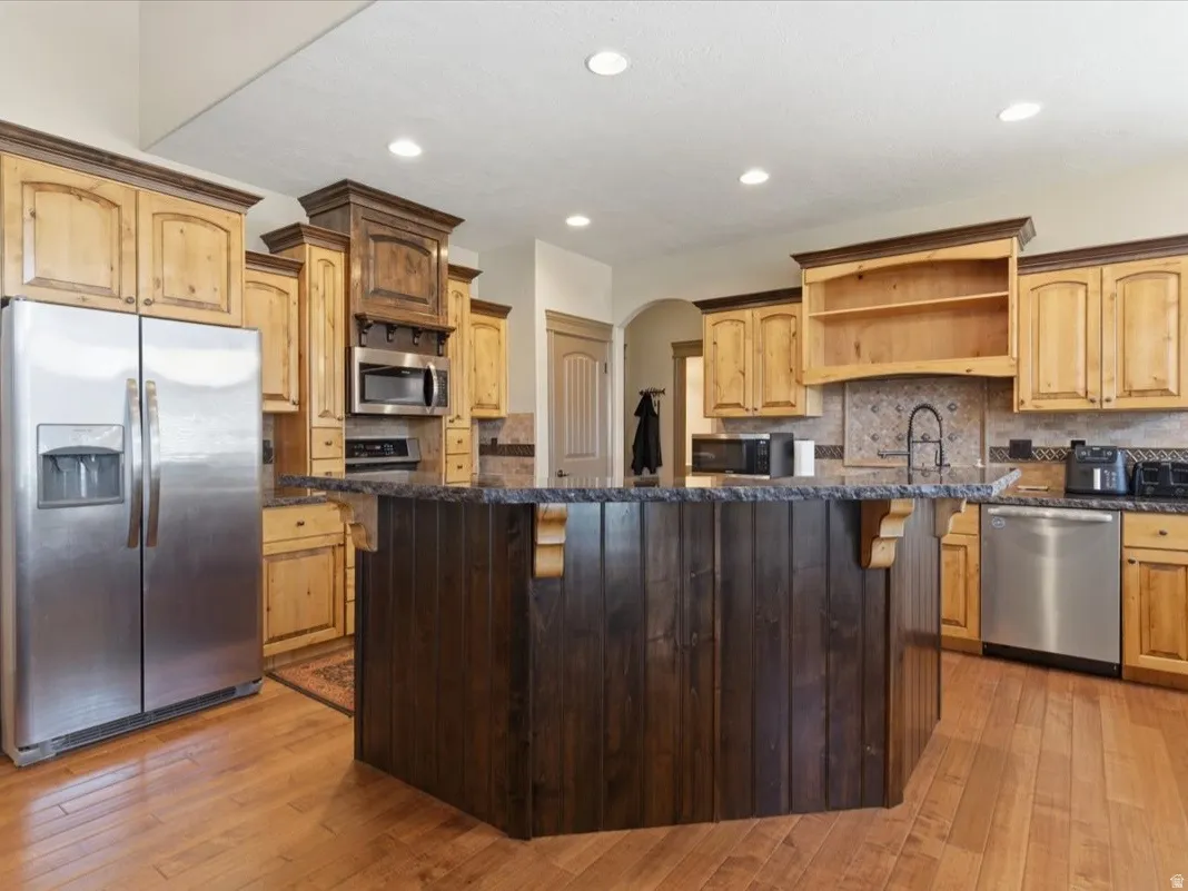 Kitchen featuring an expansive dark wood-paneled island with an integrated sink, complementing light wood-finish cabinetry, stainless steel appliances, a tiled backsplash, and wood-finish flooring
