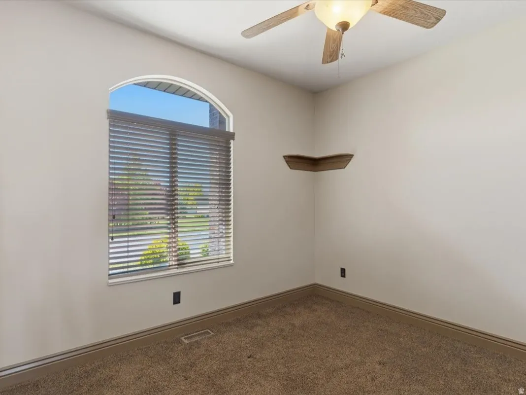 Arched window with blinds, ceiling fan, corner shelving, neutral wall color, and carpeted flooring