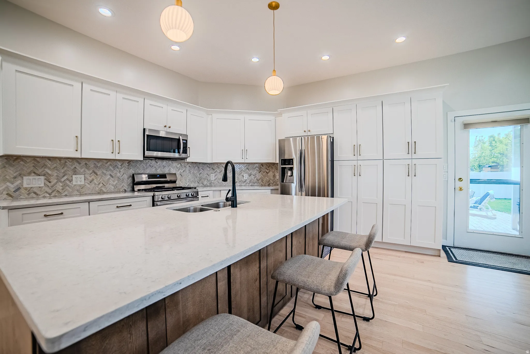 Two tone kitchen featuring light stone countertops, an island with sink, stainless steel appliances, pendant lighting, and light wood-style floors