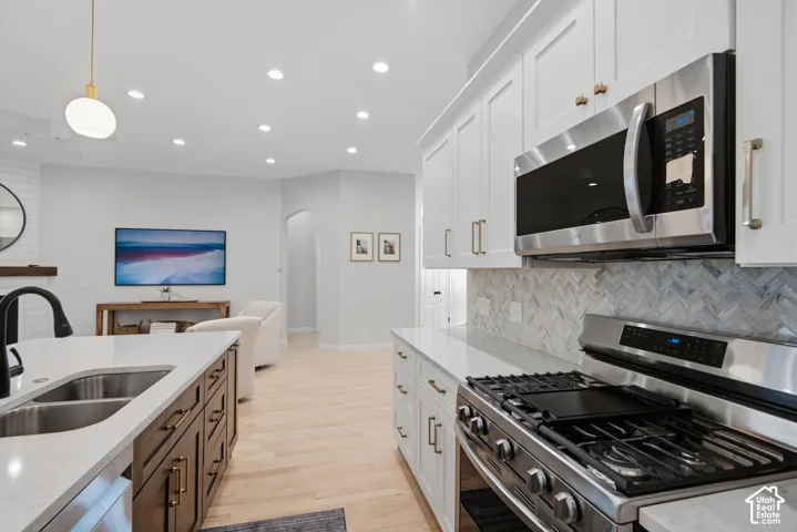 Stainless steel appliances, white cabinetry with gold-tone hardware, a herringbone pattern backsplash, light wood-finish flooring, and recessed lighting