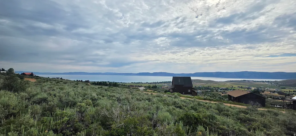 Expansive land parcel featuring natural shrubbery and established residential structures, with a prominent lake and distant mountains forming a scenic backdrop