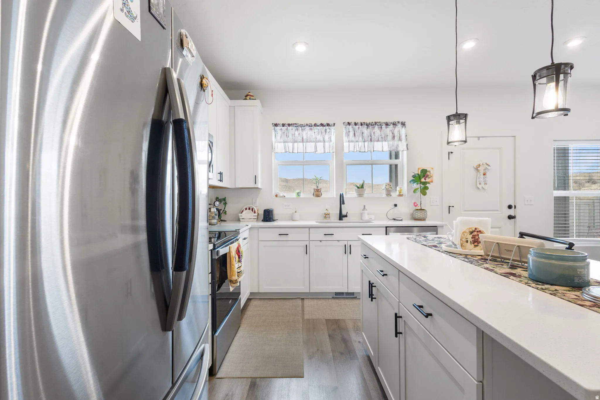 Kitchen with stainless steel appliances, white cabinets, decorative light fixtures, light wood-type flooring, and light stone counters