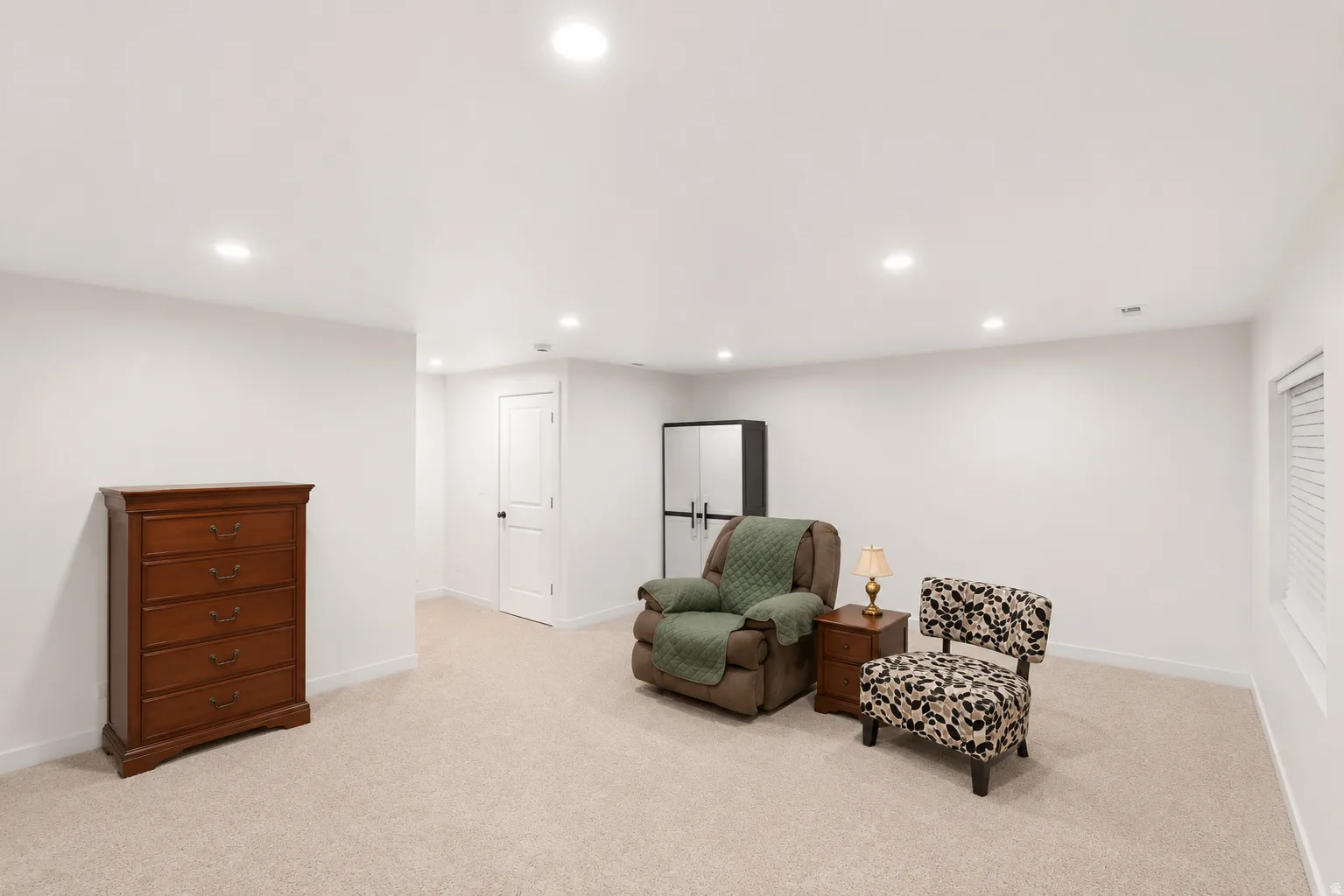 Spacious room featuring neutral carpeting and white walls, recessed lighting throughout, a white paneled interior door with dark hardware, and an egress window