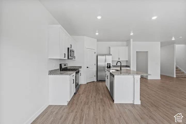 Kitchen featuring stainless steel appliances, white cabinetry, light stone counters, light wood-style floors, and a center island with sink