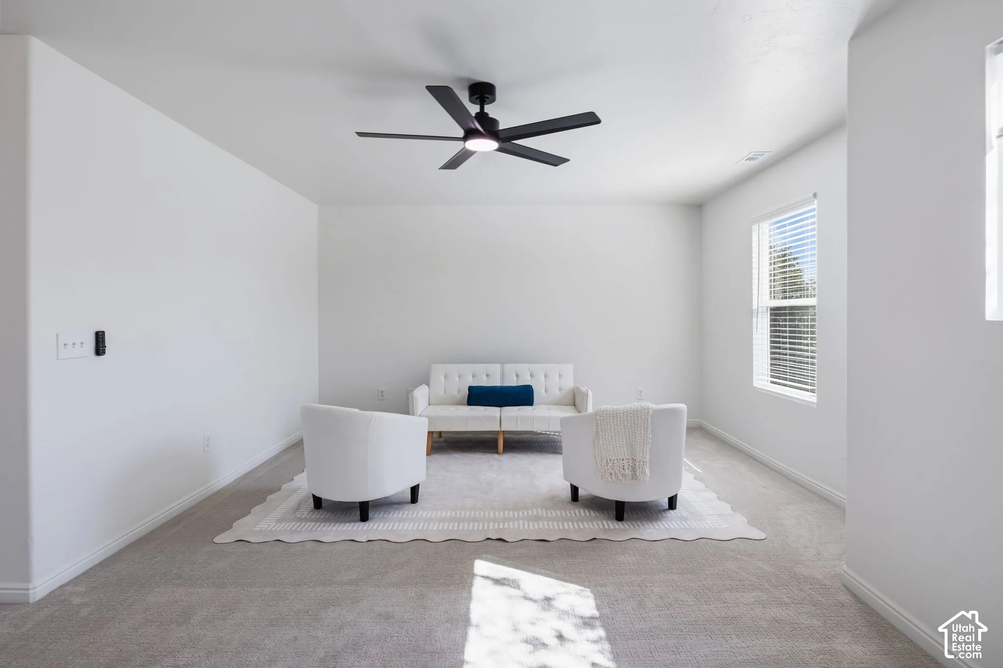 Spacious room with plush carpet flooring, a modern black ceiling fan, and ample natural light from a window with blinds