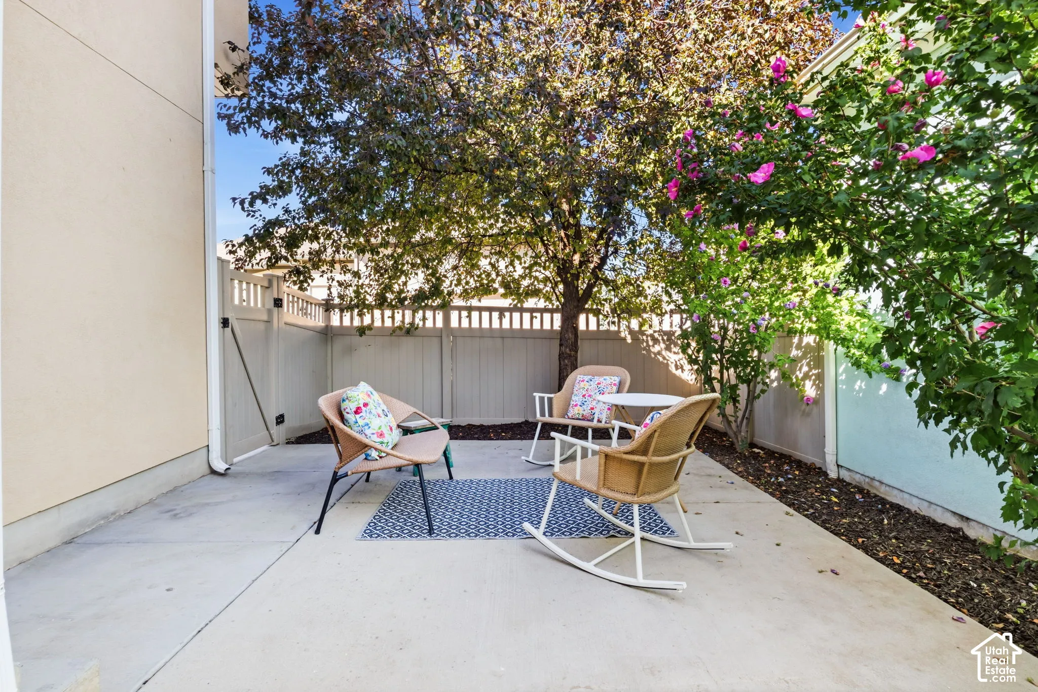 Concrete patio area with a privacy fence, mature shade tree, and flowering shrubbery