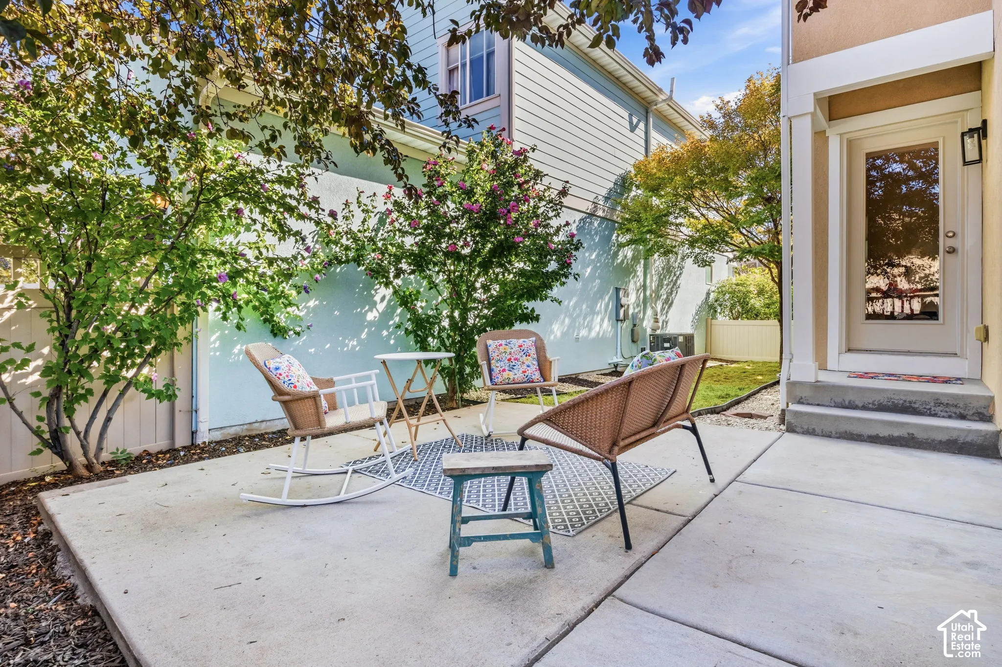 Concrete patio area with a privacy fence, mature landscaping, and an exterior door with a wall-mounted light fixture