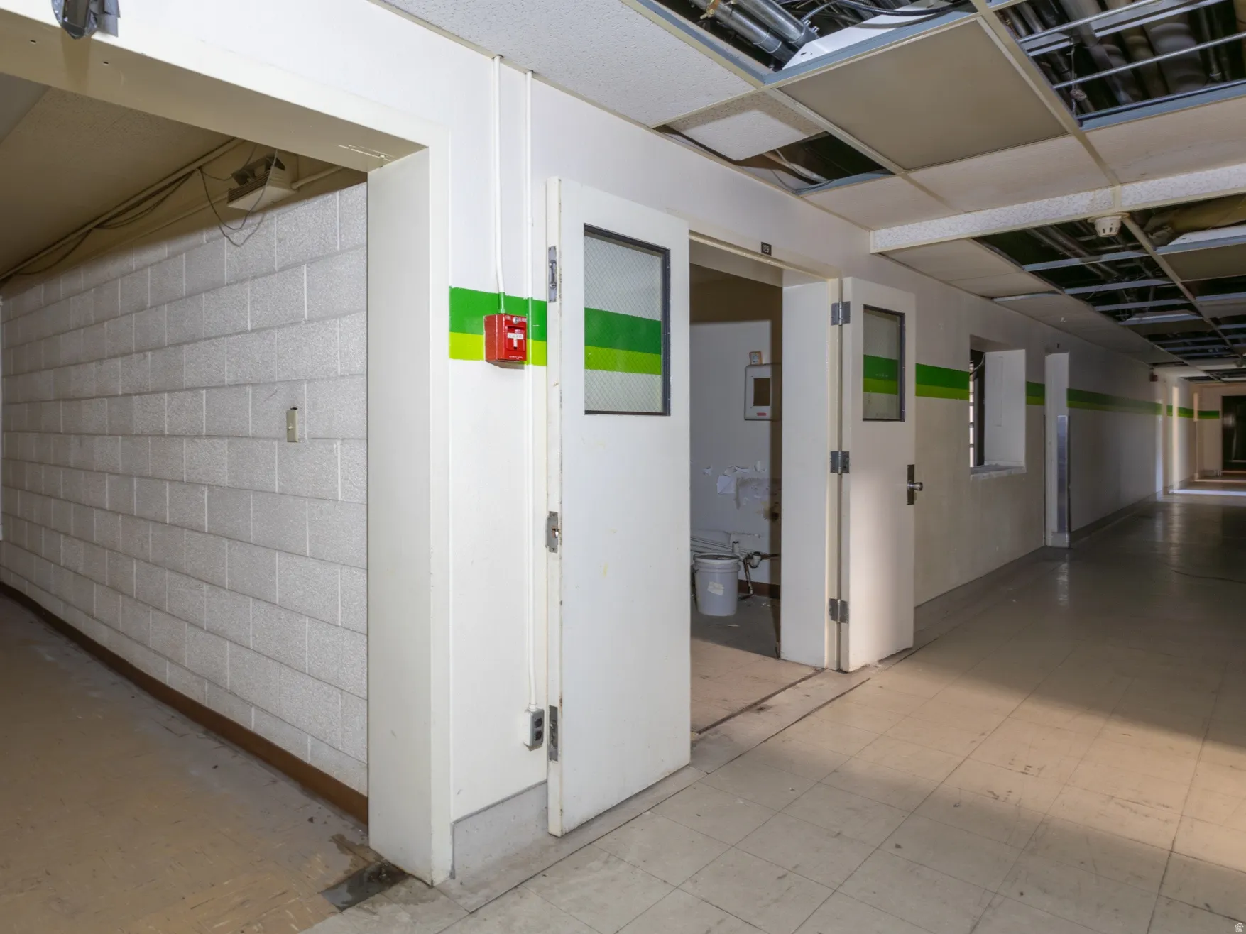 Corridor featuring a concrete block wall, multiple doorways with vision panels, and a drop ceiling with exposed infrastructure