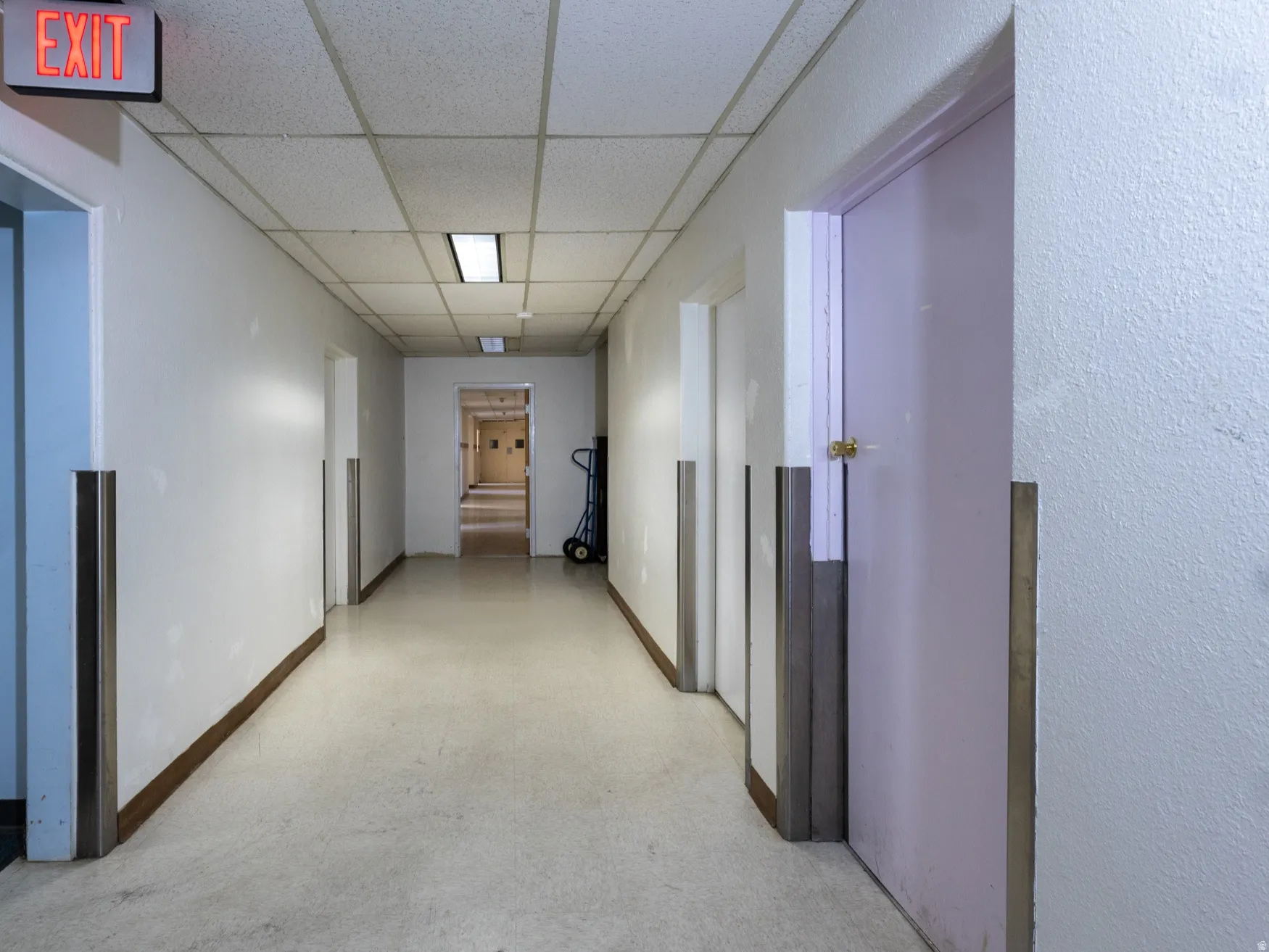 Long hallway with light-toned flooring, drop ceiling panels, and recessed fluorescent lighting fixtures