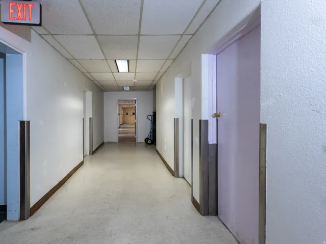 Long hallway with light-toned flooring, drop ceiling panels, and recessed fluorescent lighting fixtures