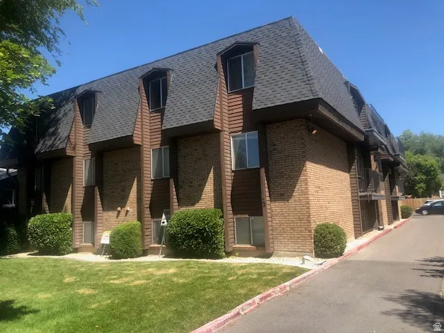 Multi-story building featuring brick and vertical wood-finish siding, dormer windows, a dark shingle roof, and a grassy lawn with manicured shrubs