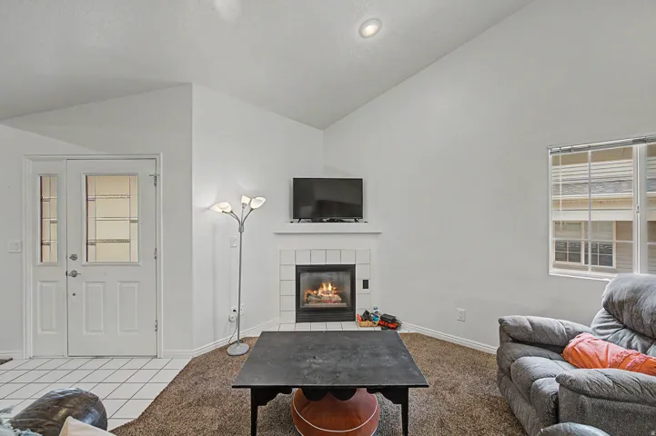 Living area featuring a corner fireplace with a white tiled surround and mantel, vaulted ceiling, and a large window with horizontal blinds