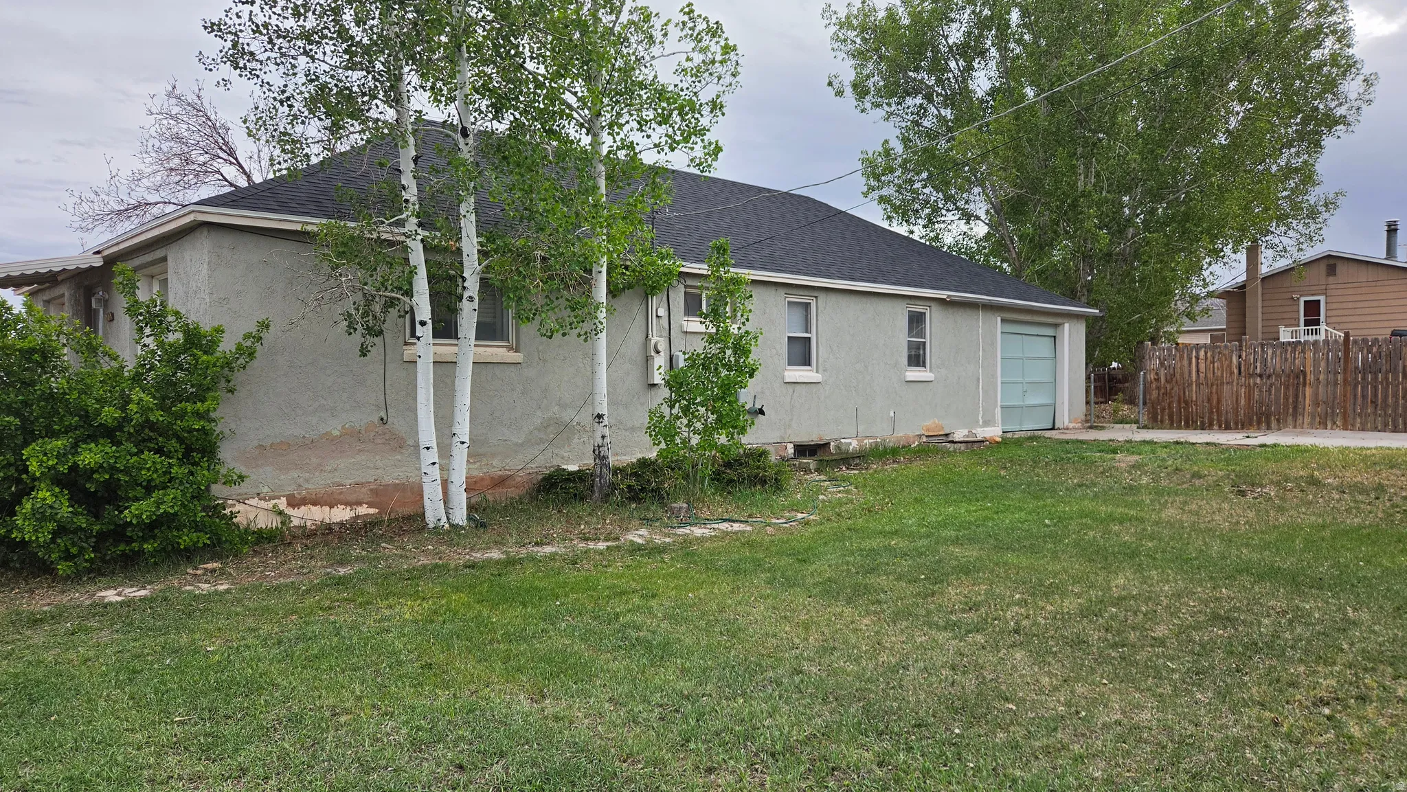 Stucco exterior residence featuring a dark shingle roof, attached garage with a light blue door, mature landscaping, and partial fencing