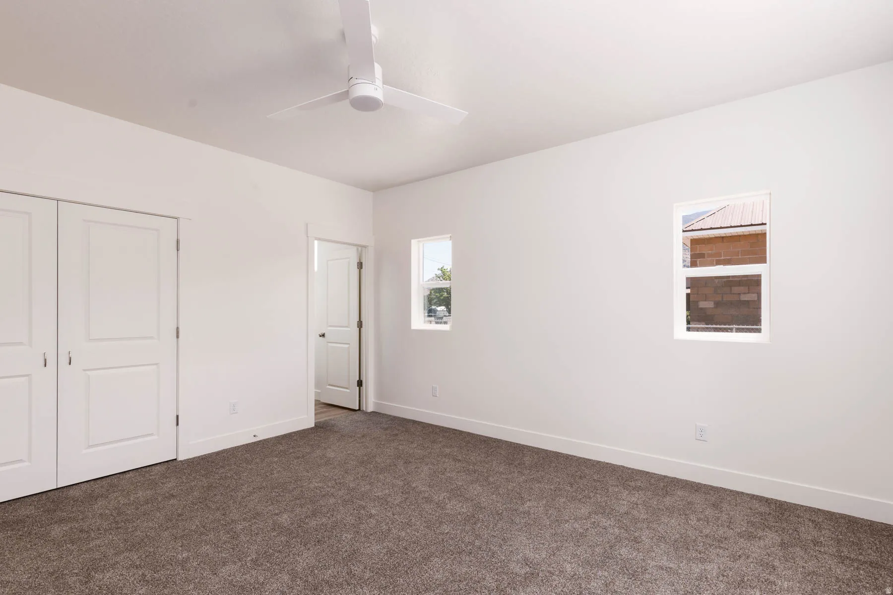 Interior room featuring a white ceiling fan, two windows providing natural light, white walls, and dark carpeting
