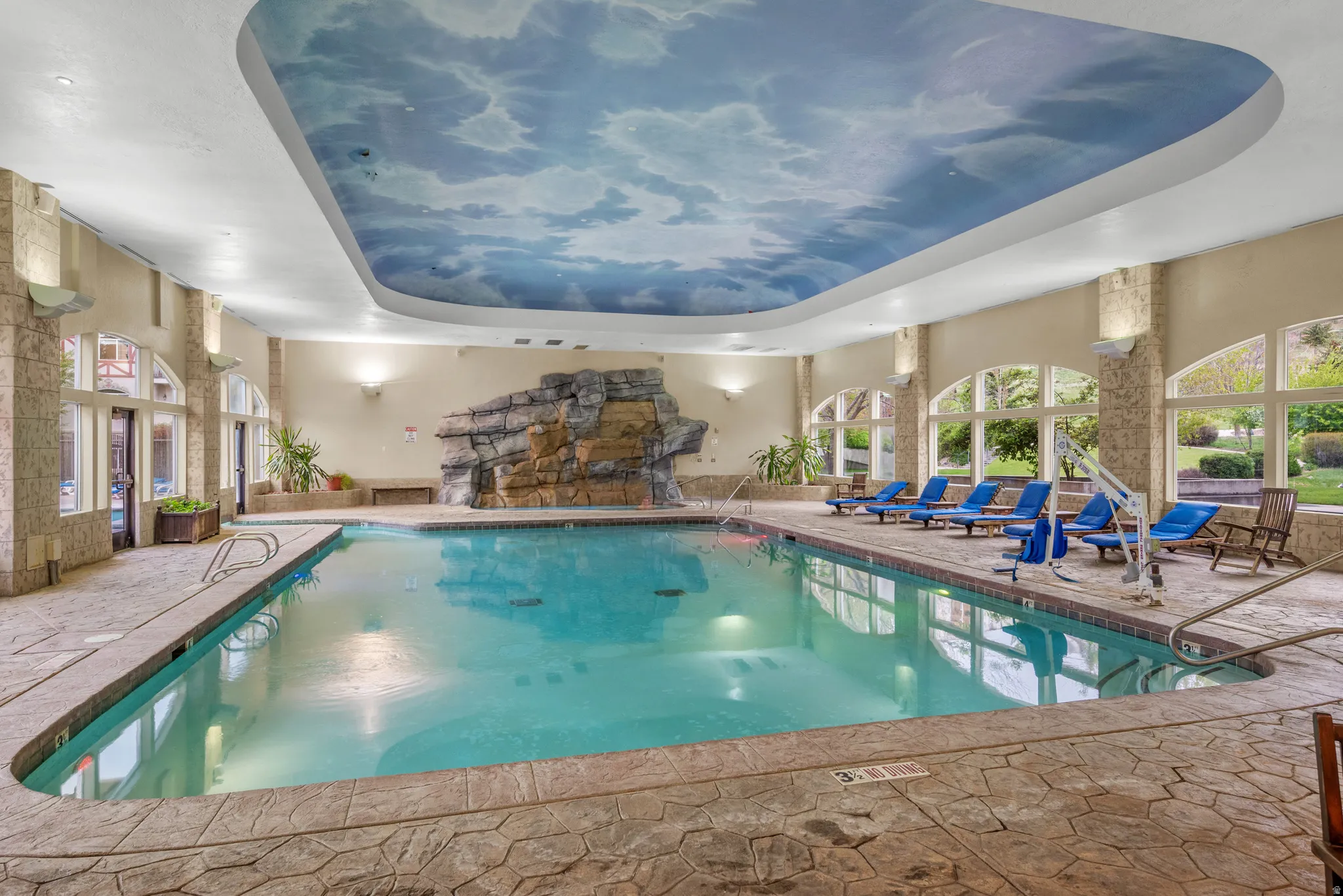 Indoor swimming pool featuring a decorative rock waterfall, a cloud-mural ceiling, and arched windows