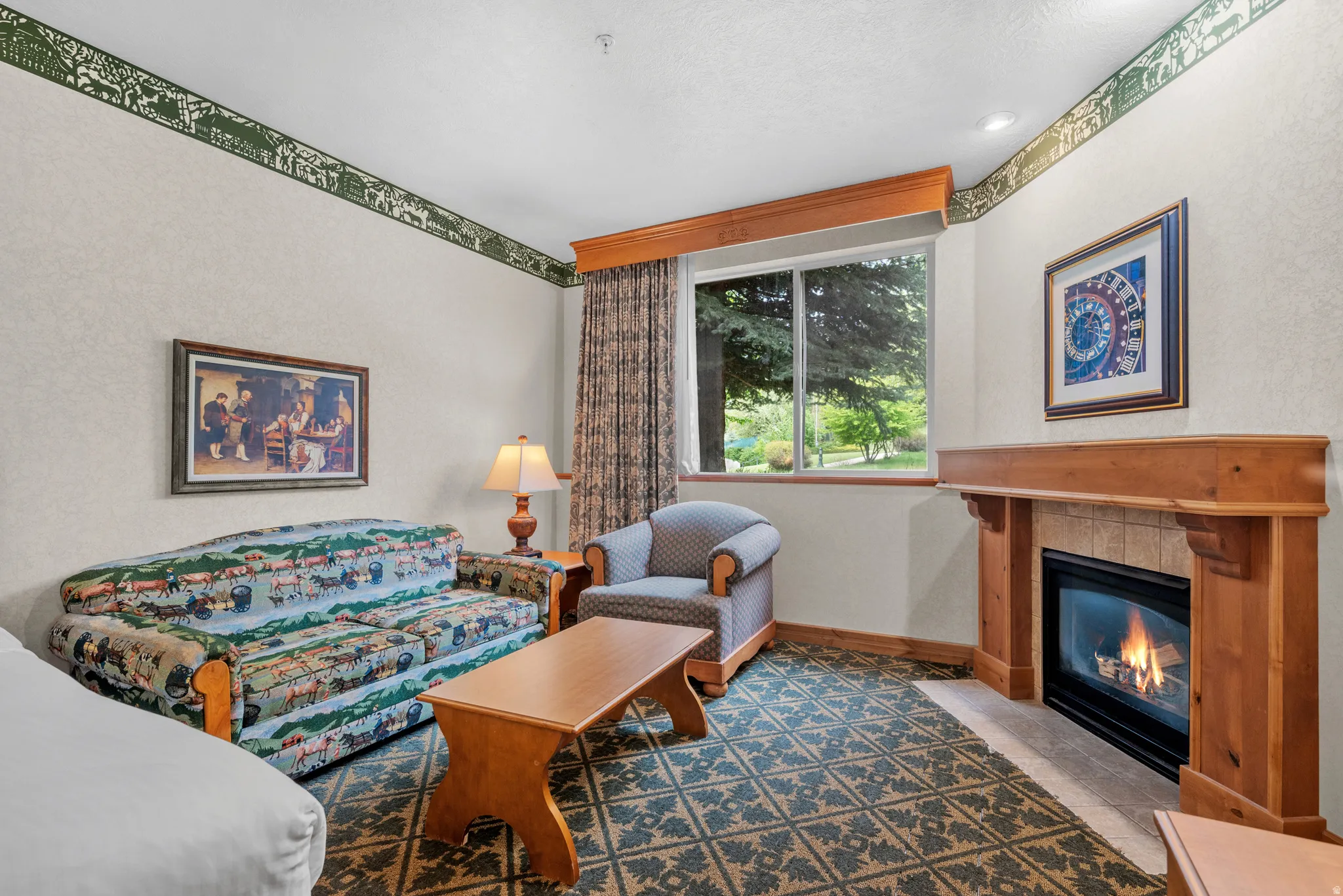 Living area featuring a gas fireplace with a wood mantle and tile surround, large window with outdoor greenery, patterned carpet flooring, and a decorative wallpaper border