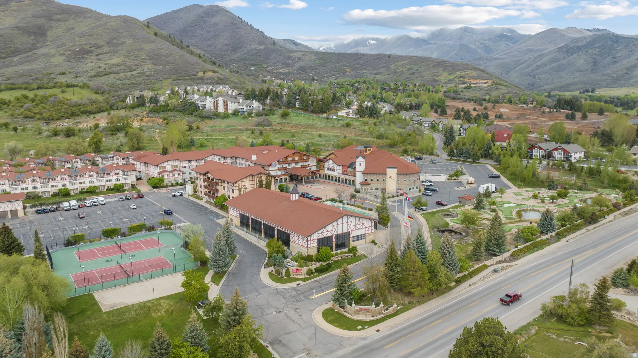 Aerial overview showcasing a multi-building property featuring prominent terra cotta tile roofs, extensive parking, multiple tennis courts, and a miniature golf course, all set against a backdrop of mountainous terrain