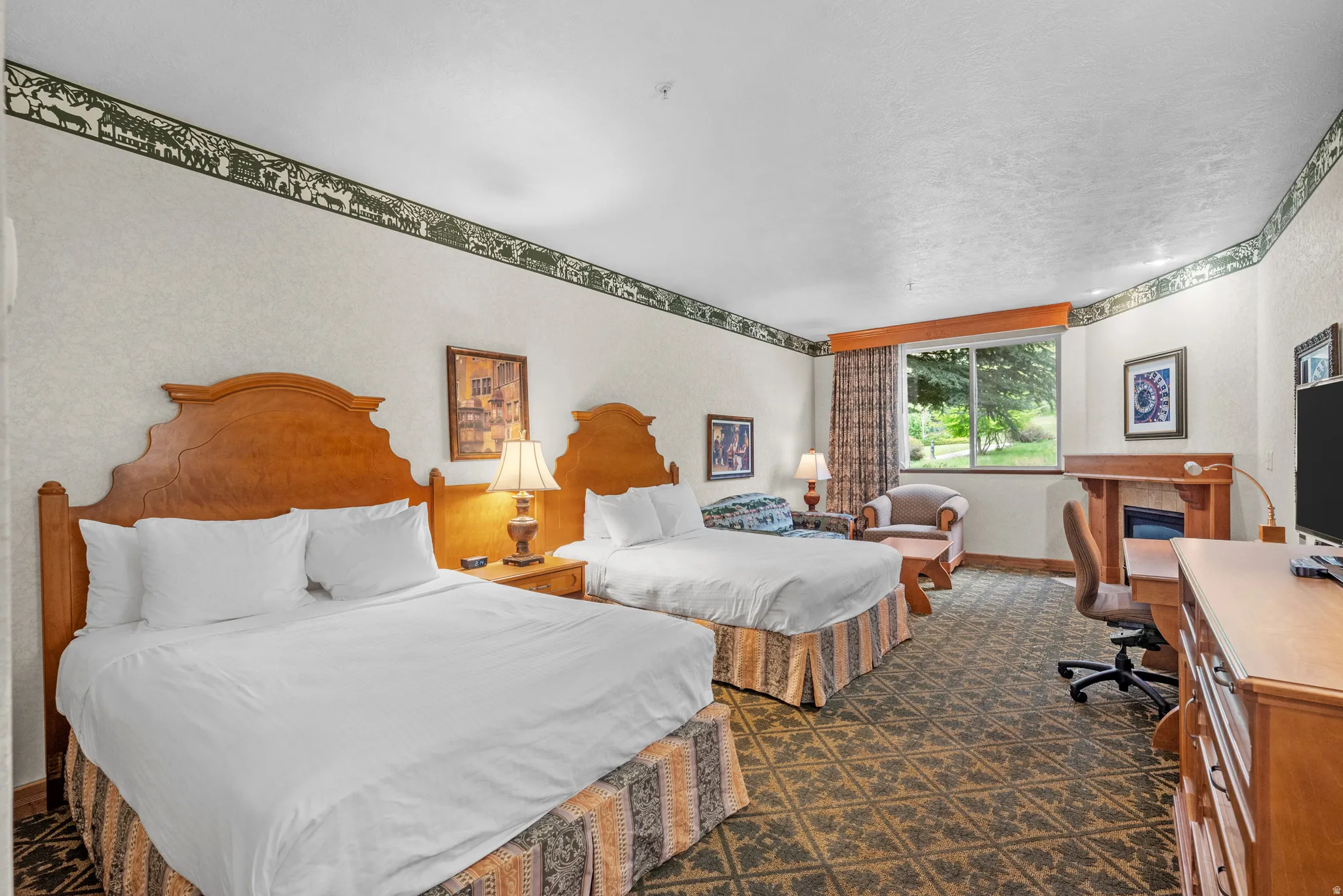 Spacious interior featuring two wood-finish headboards, patterned carpeting, a large window, a built-in electric fireplace, and a dedicated desk area