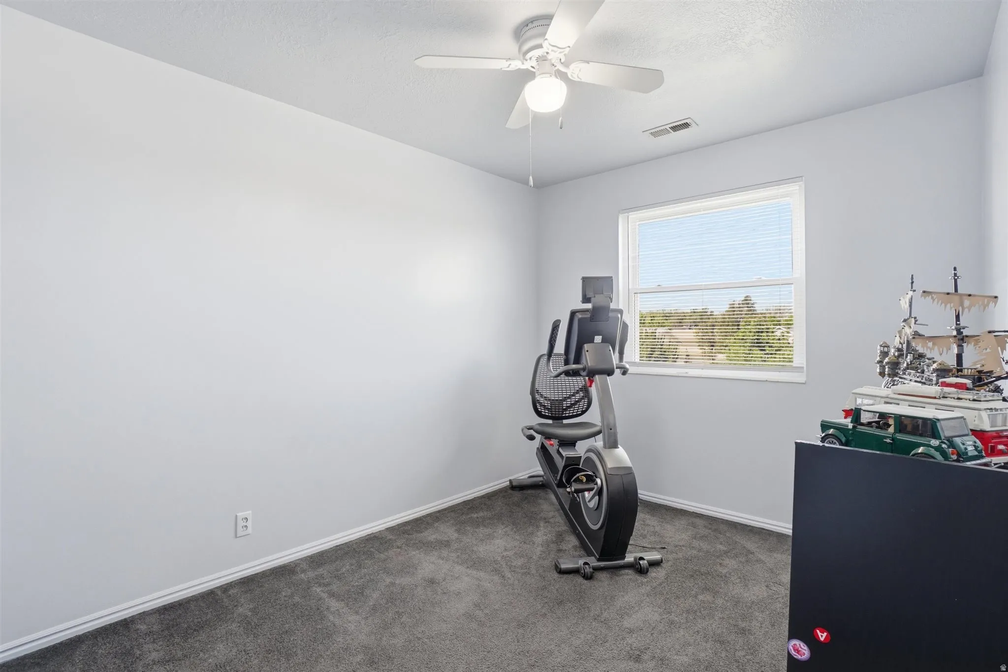Bright room featuring a window with blinds, ceiling fan with light, gray wall-to-wall carpeting, and white baseboards