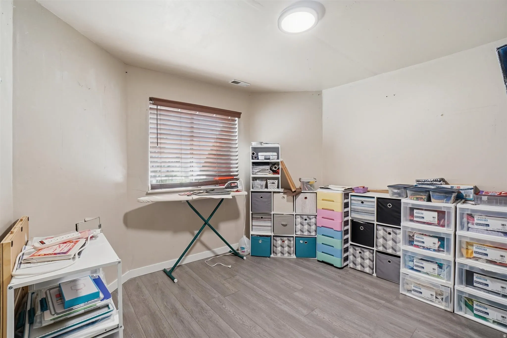 Spacious room featuring wood-finish flooring, a window with blinds, and a flush-mount ceiling light