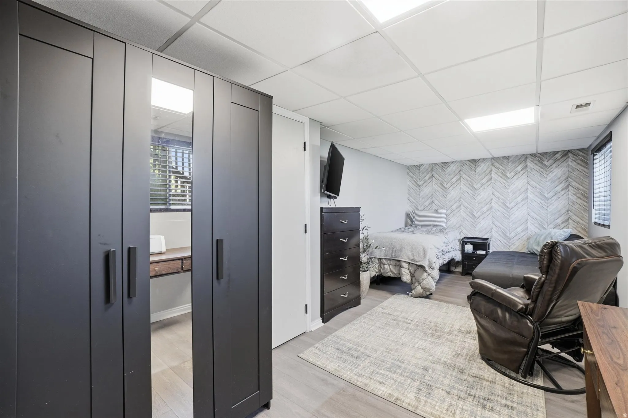 Spacious room featuring light wood-finish flooring, a decorative accent wall, white walls, and a dropped ceiling with integrated lighting