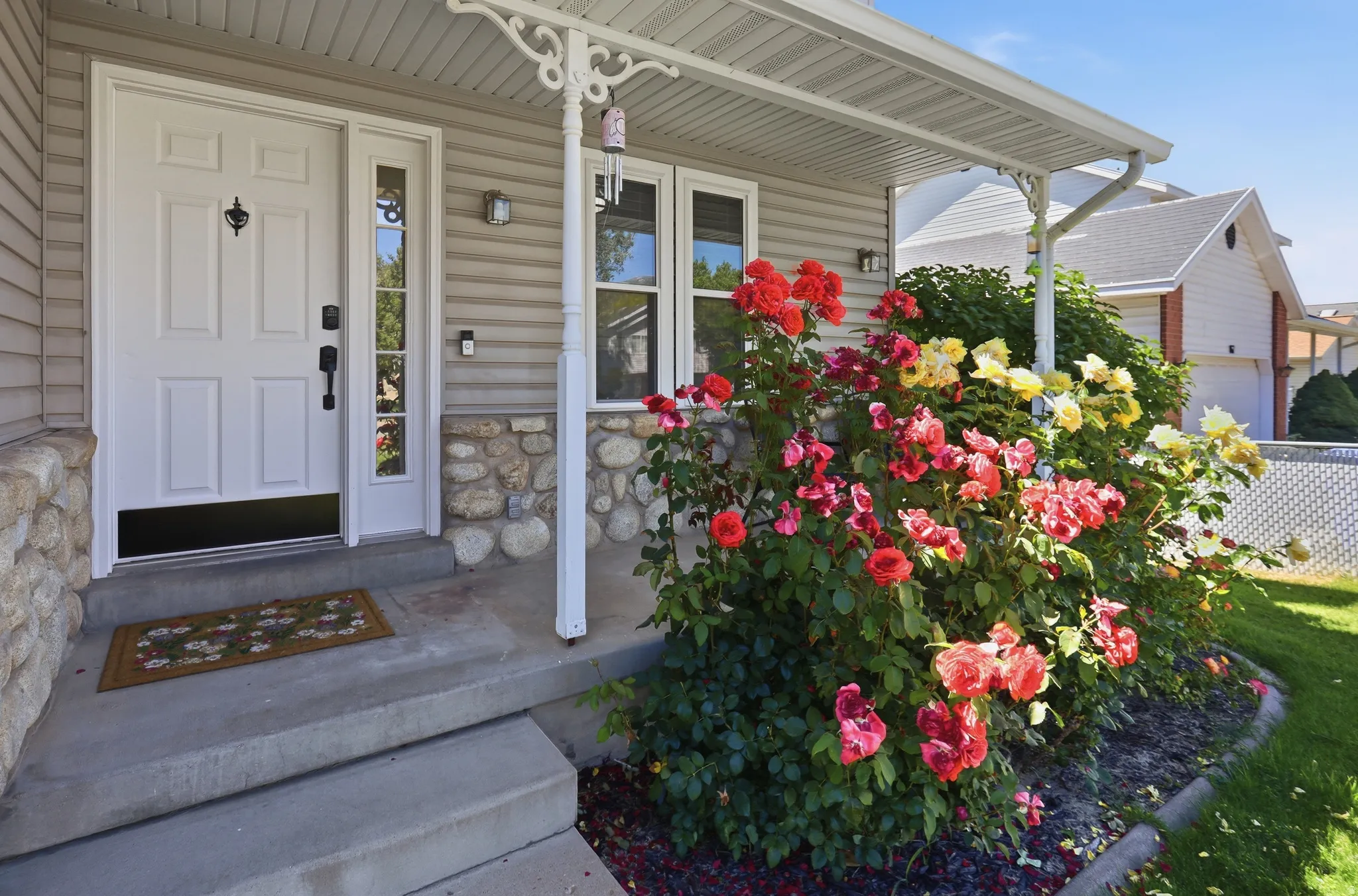 Inviting front entry featuring a paneled door with a sidelight, covered porch, concrete steps, stone veneer accents, and vibrant rose bushes