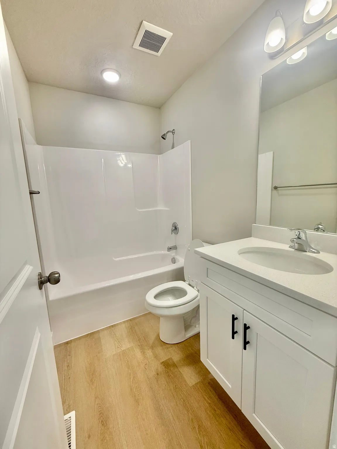 Bathroom featuring a white vanity with a white countertop and black hardware, a shower-tub combination with a chrome showerhead and faucet, and wood-finish flooring