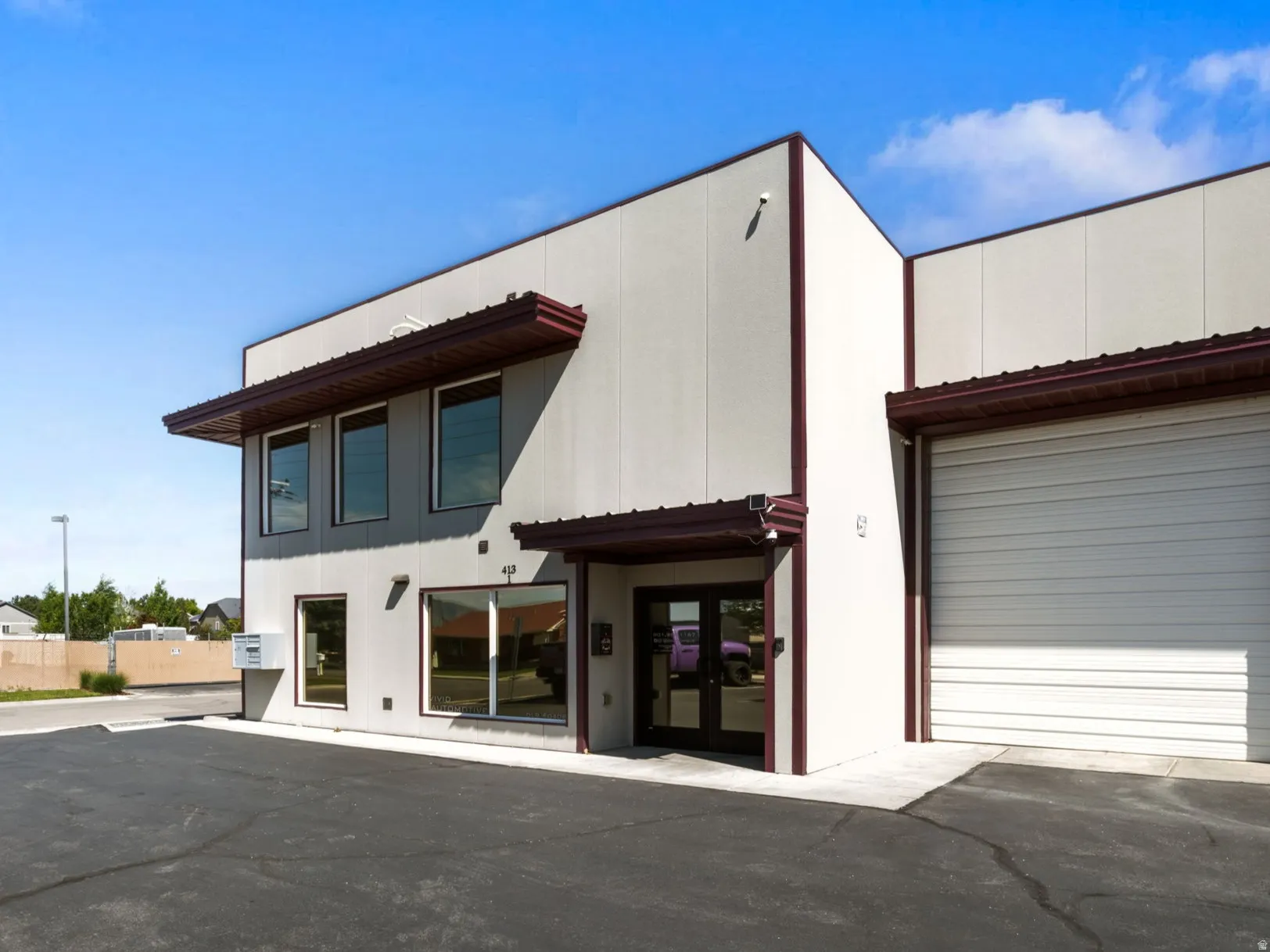 Modern commercial building featuring a light-colored stucco exterior, dark brown trim, multiple large windows, a prominent roll-up door, and an asphalt parking lot
