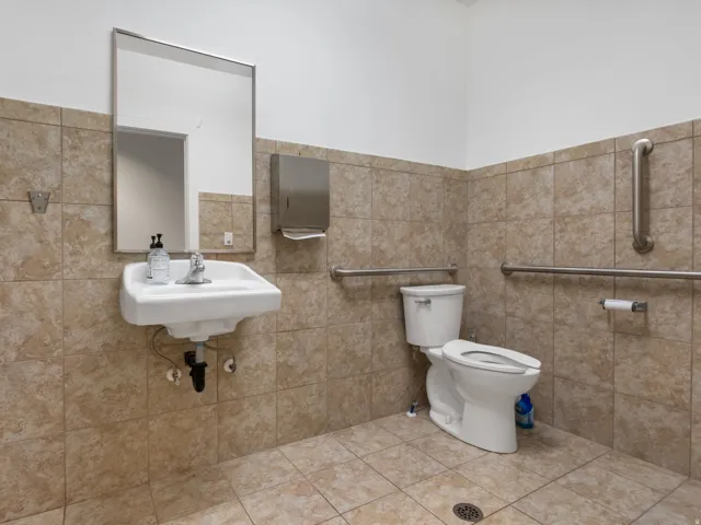 Restroom featuring floor-to-ceiling ceramic tile, a wall-mounted sink with a chrome faucet, a white toilet, and multiple stainless steel grab bars