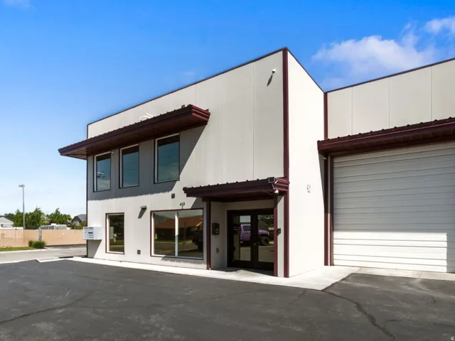 Modern commercial building featuring a light-colored stucco exterior, dark brown trim, multiple large windows, a prominent roll-up door, and an asphalt parking lot