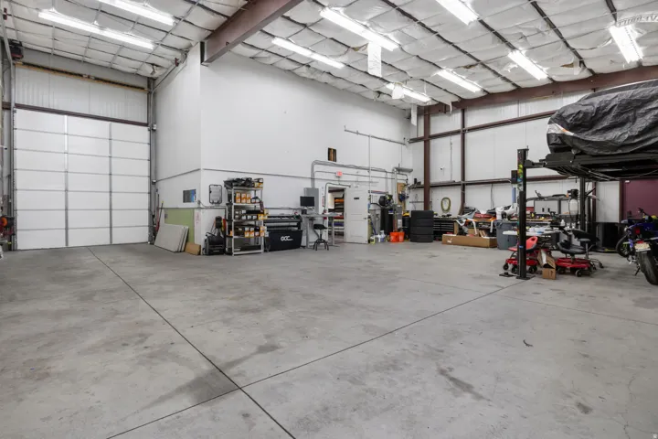 Spacious warehouse interior featuring a large roll-up door, high ceilings with exposed metal beams, concrete flooring, and ample overhead fluorescent lighting