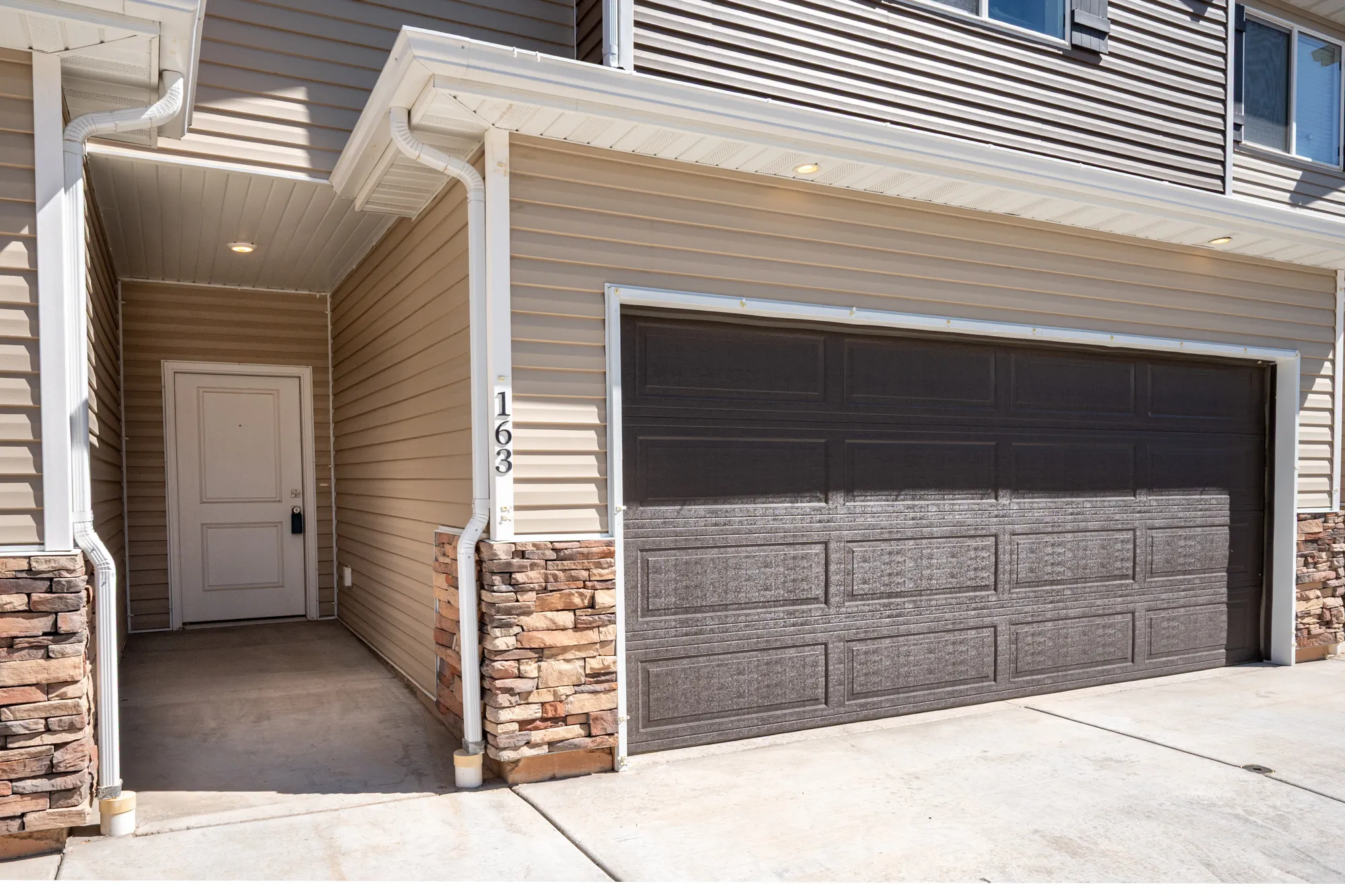 Attached garage featuring a textured brown door, stone veneer accents, and tan siding