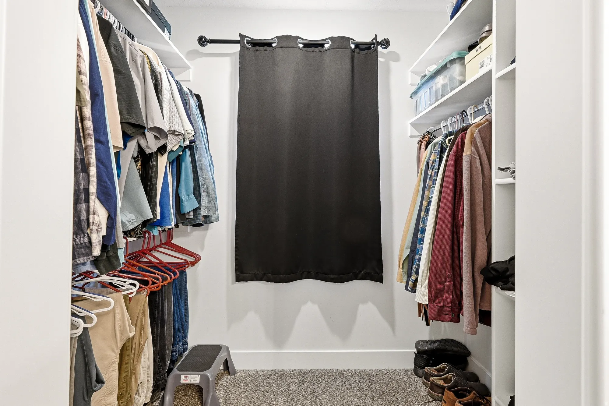 Walk-in closet featuring built-in shelving, hanging rods, light gray carpeting, and a window with a dark curtain