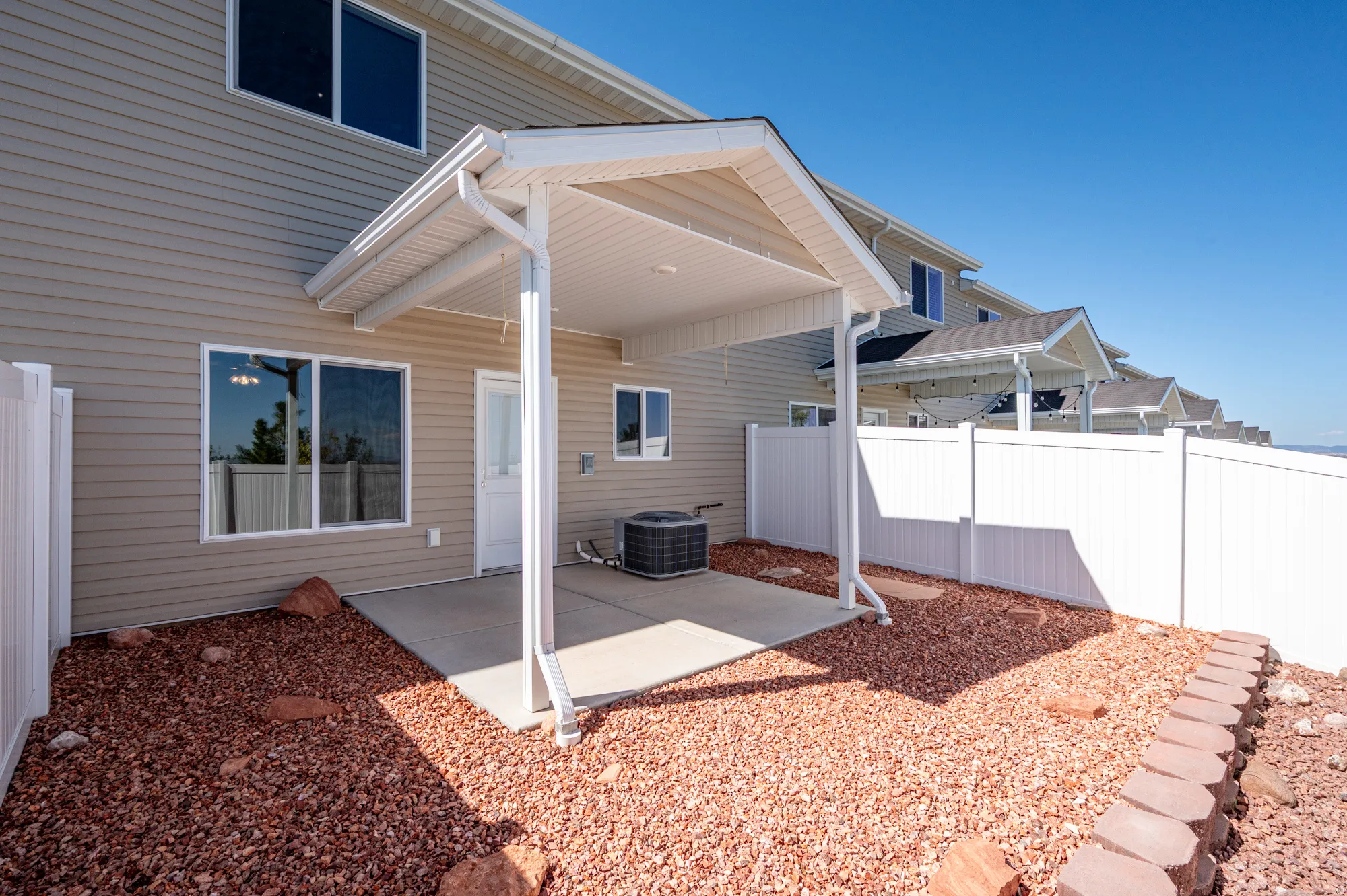 Rear patio area featuring a covered concrete pad, light beige horizontal siding, white vinyl fencing, and surrounding reddish-brown gravel landscaping