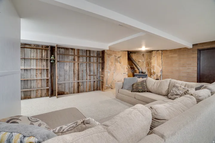 Finished lower level featuring a wood-burning stove with a stone surround, wood-paneled walls with built-in shelving, carpet flooring, and exposed ceiling beams