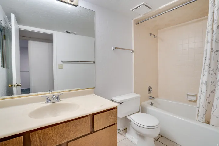 Bathroom featuring a wood-finish vanity with an integrated sink and chrome faucet, a large wall-mounted mirror, a toilet, and a combined bathtub and shower with light-toned wall tile