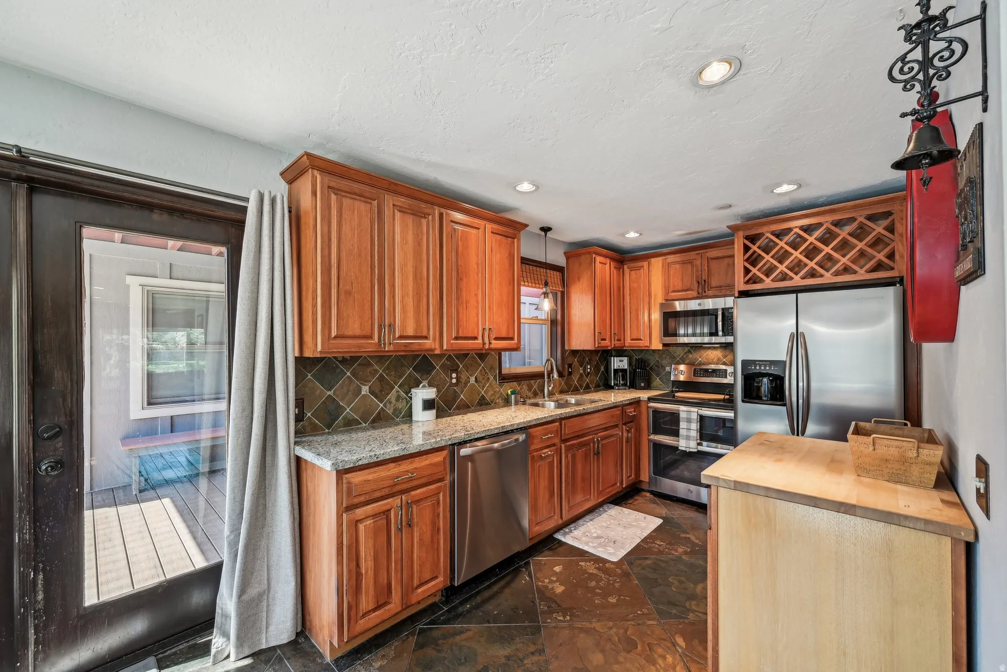 Kitchen featuring stainless steel appliances, decorative light fixtures, wood finish cabinetry, tasteful backsplash, and dark stone finish flooring