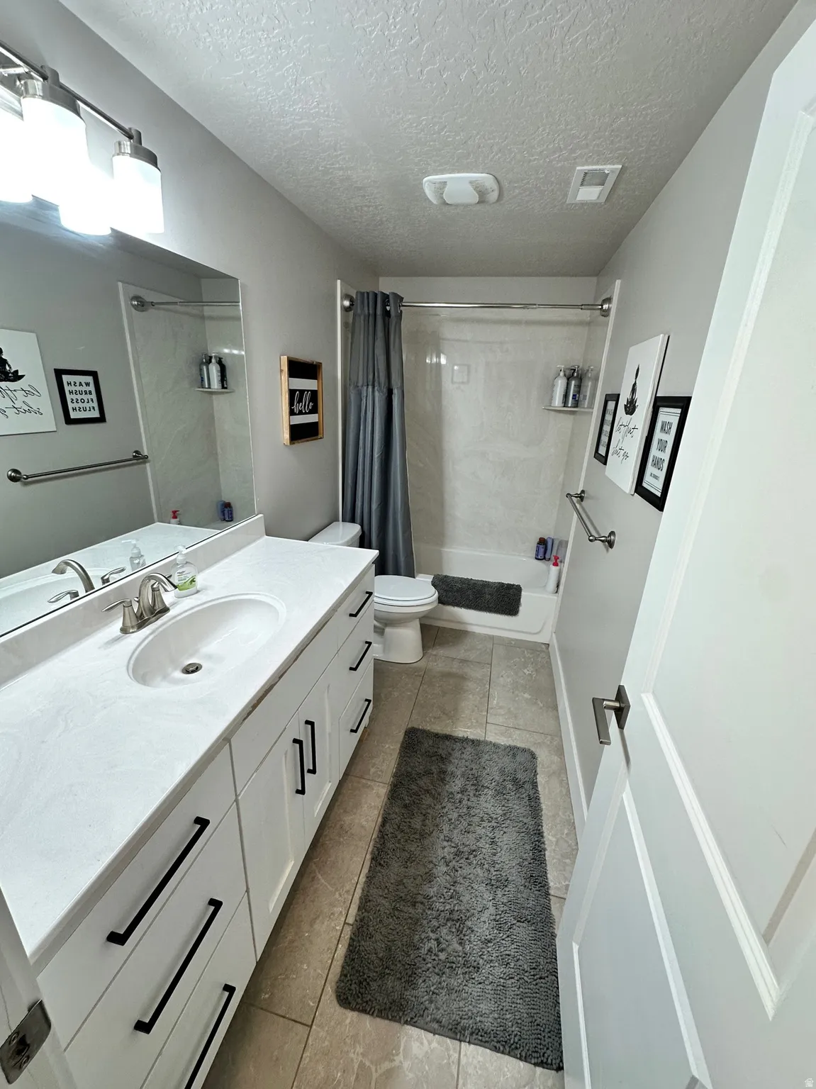 Bathroom featuring a single vanity with a white countertop and undermount sink, white cabinetry with black hardware, a shower-tub combination with a white surround, and tile flooring