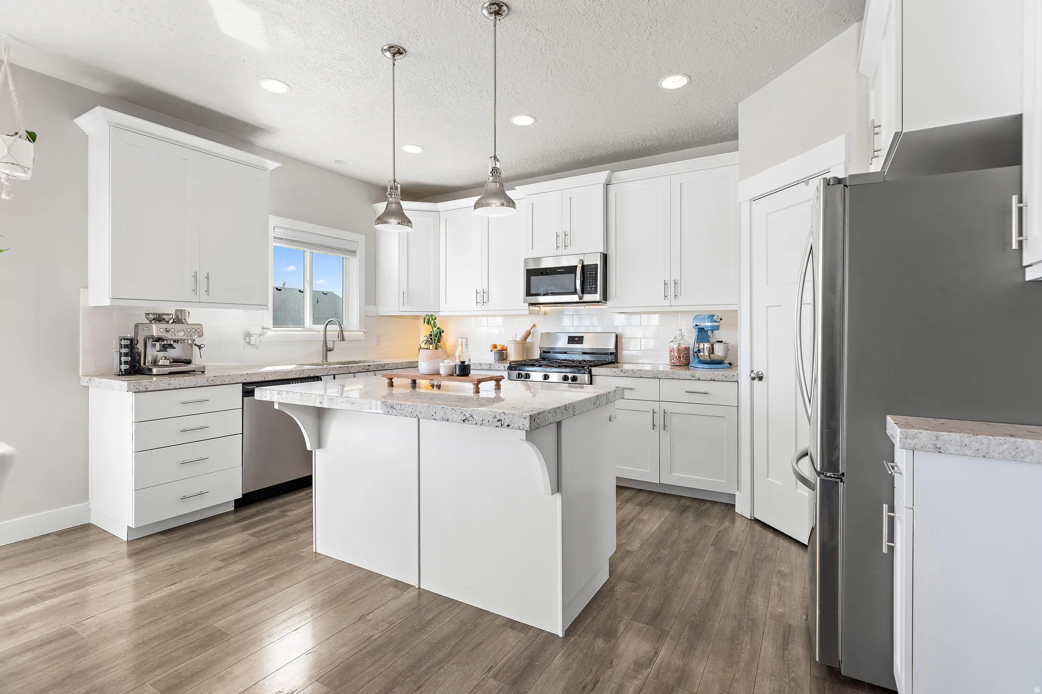 Modern kitchen featuring white cabinetry, light-colored countertops, stainless steel appliances, a center island with pendant lighting, and wood-finish flooring