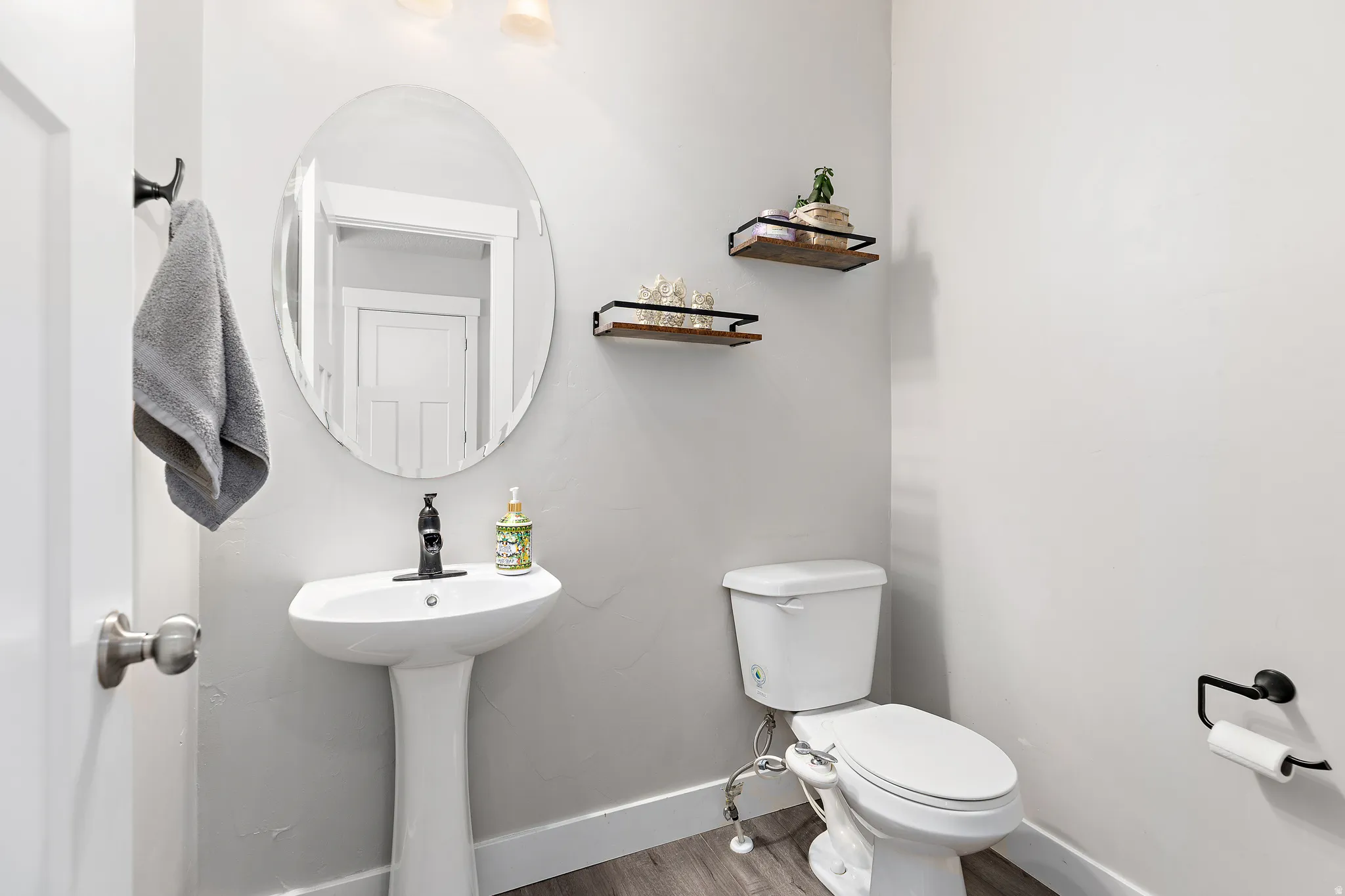 Pedestal sink with a black faucet, oval mirror, and wall-mounted shelving