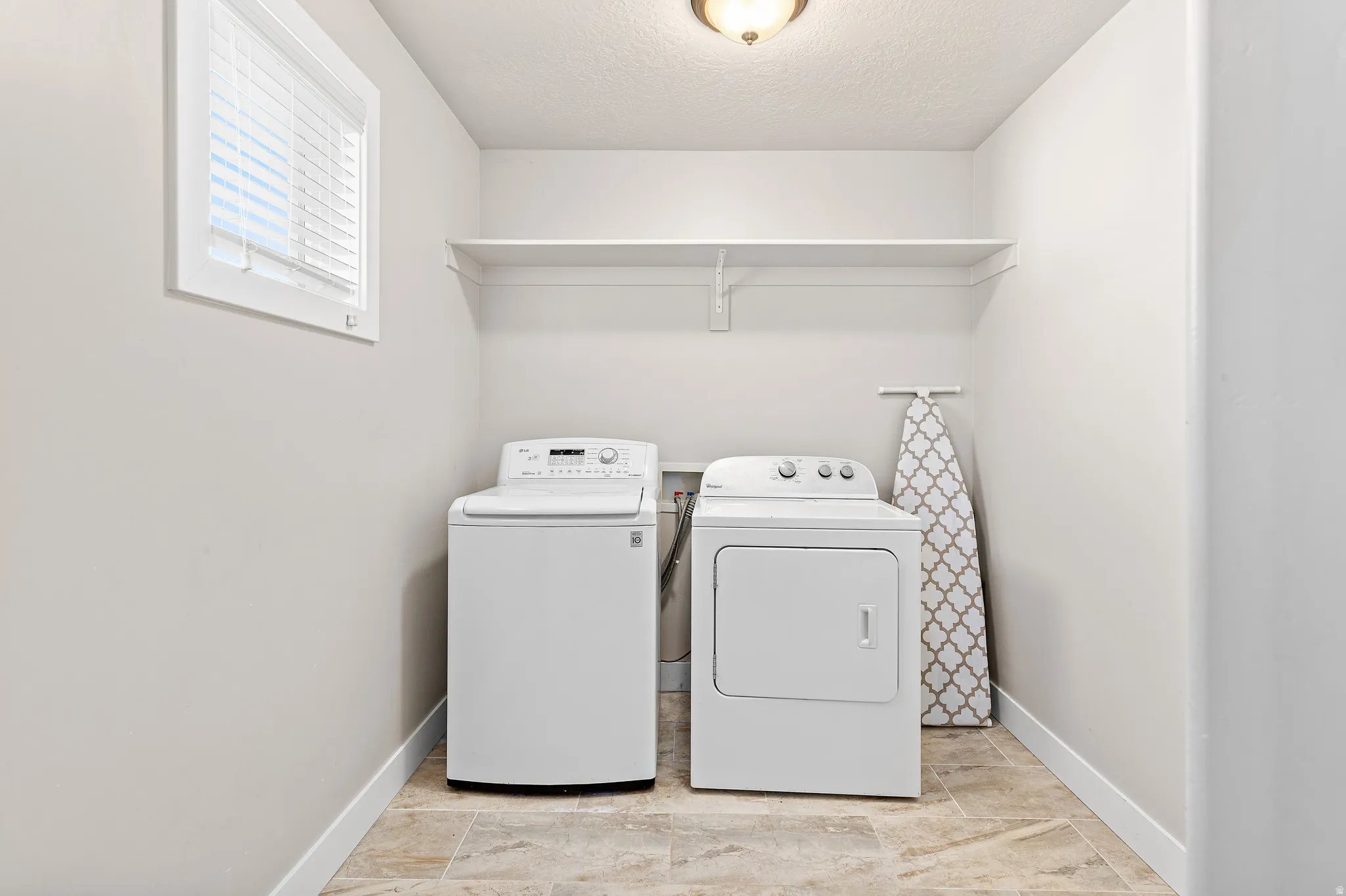 Dedicated utility room featuring light neutral wall tones, a single window with blinds, a built-in overhead shelf, and tiled flooring
